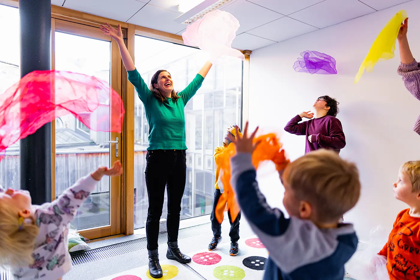 Children's music class, the teacher and the children are throwing colourful scarves in the air, everyone is smiling and laughing.